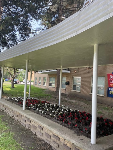 A brick building with a white metal awning supported by columns, with red and white flowers planted in beds along the front.