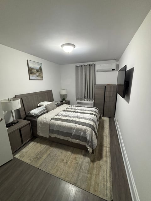 Modern bedroom with dark wood bed, striped bedding, nightstand, flat-screen TV, and gray curtains on white walls