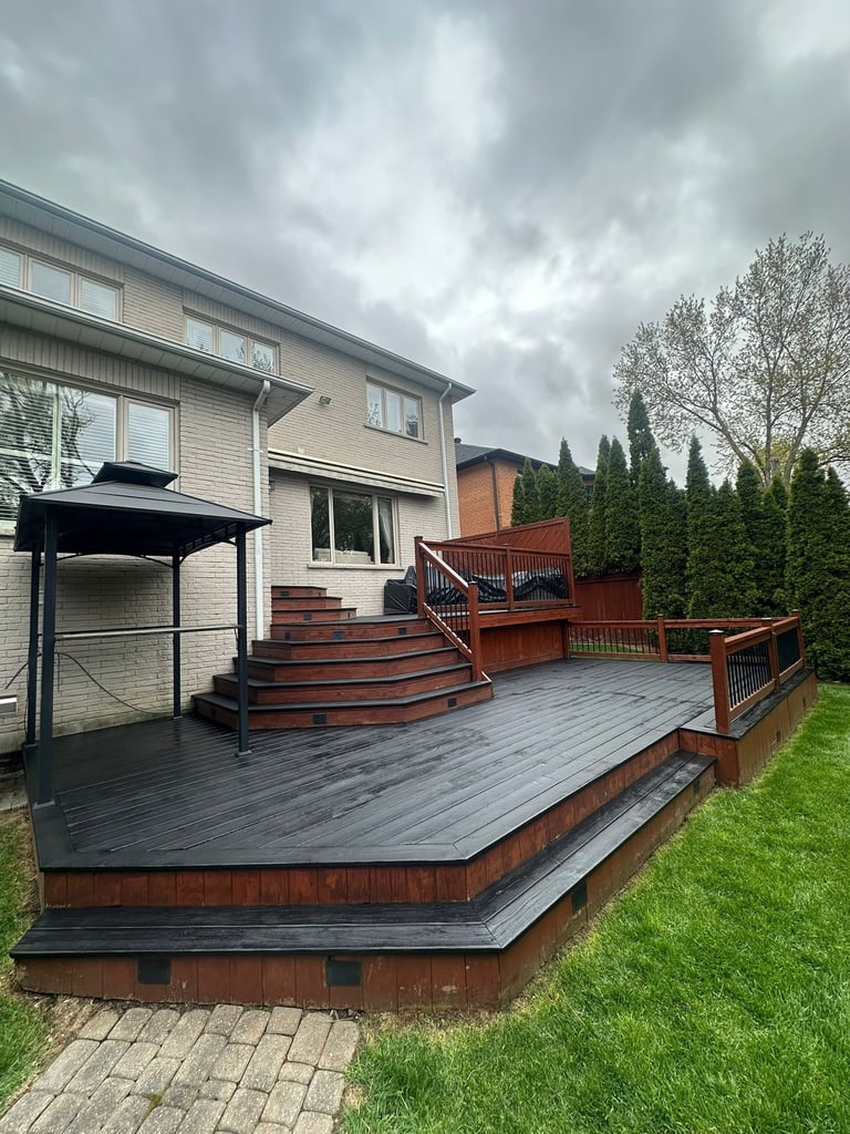 Two-story house with wood and composite deck, red stairs, black canopy, green lawn, and evergreen trees on overcast day