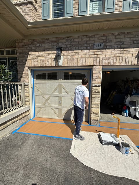 Person standing in garage driveway with blue tape marking floor, protective coverings in place for renovation work on brick home