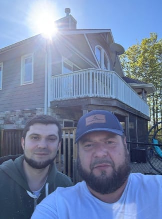 Two men taking a selfie in front of a raised brick house with a deck on a sunny day