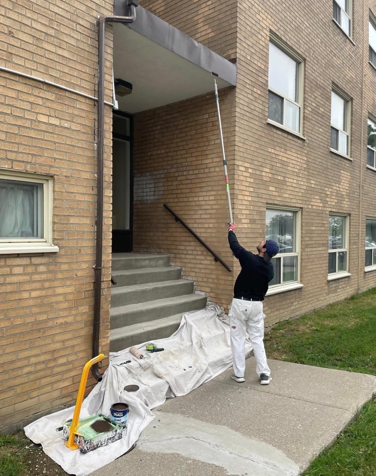 Person in black shirt and white pants cleaning building exterior with long pole reaching toward roof overhang