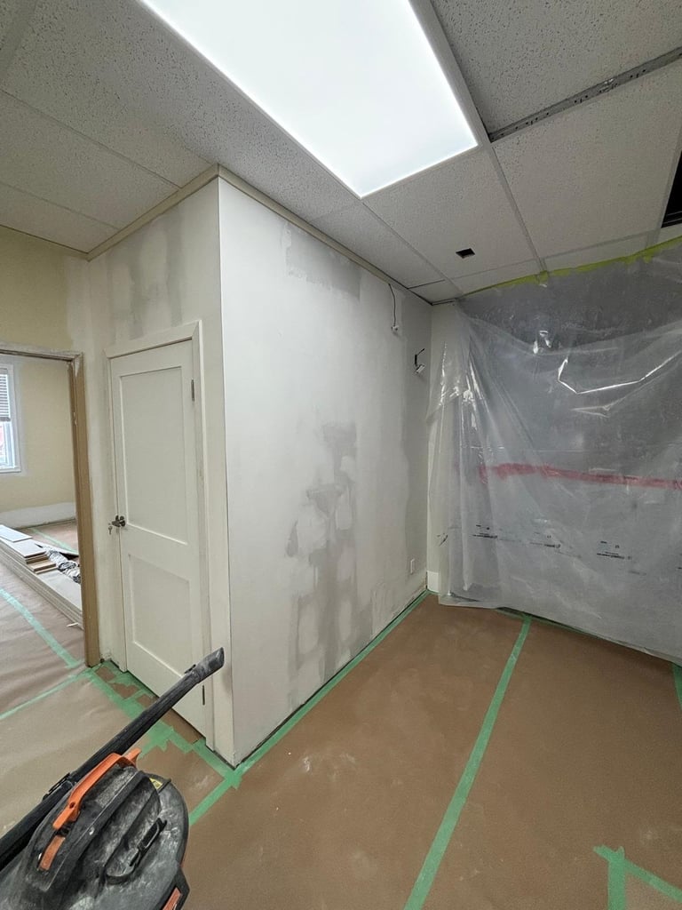 Interior room under renovation with plastic sheeting on walls, drop cloth on floor, and green tape marking, with white door visible.