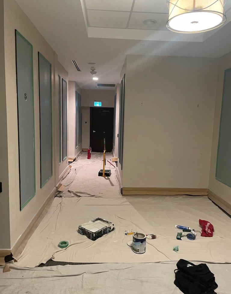 Commercial hallway under renovation with white walls, drop cloths, paint cans, tools, and construction materials scattered on the floor
