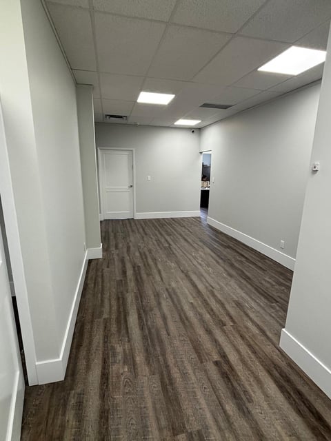 Empty modern office corridor with white walls, recessed ceiling lights, dark wood-grain vinyl flooring, and white trim