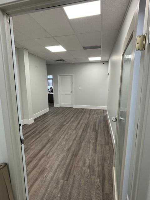 Empty office hallway with wood-look flooring, white walls, ceiling lights, and white doors on either side