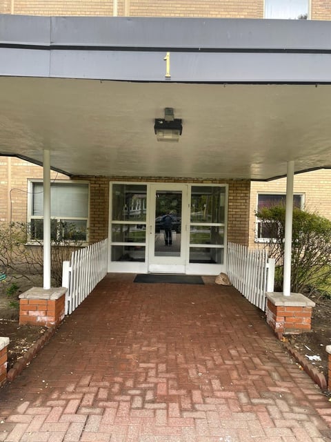 Brick building entrance with covered overhang, white railings, glass doors, and herringbone patterned brick walkway