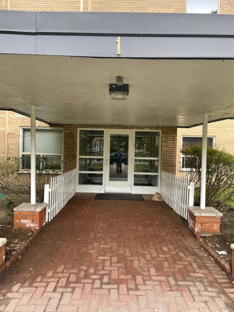 Brick building entrance with covered overhang, white railings, glass doors, and herringbone patterned brick walkway