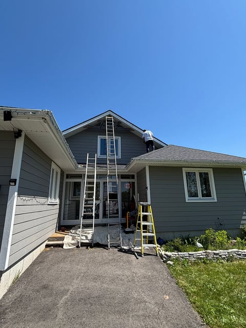 Two-story gray house under clear blue sky with ladder and yellow stepladder leaning against the garage entrance during renovation or maintenance work.